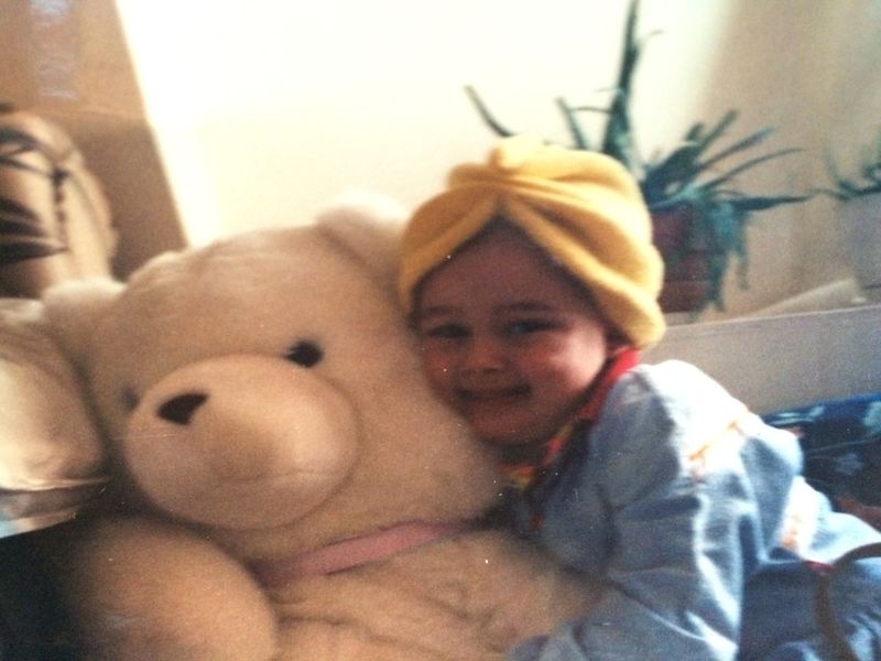 Danielle as a child in a yellow head wrap, hugging a teddy bear